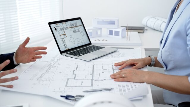 Architects and client discussing new house design, reviewing construction blueprints and digital plans on laptop for real estate project development