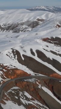 Drone footage of Chyyyrchyk Pass with winding mountain road, snow covered ridgelines and exposed red rock slopes, remote highlands landscape in Kyrgyzstan