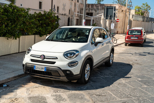 Barletta, Italy - Aug 1, 2026: White Fiat 500X crossover parked on a stone paved street with a red car behind and a hedge topped wall in the old town
