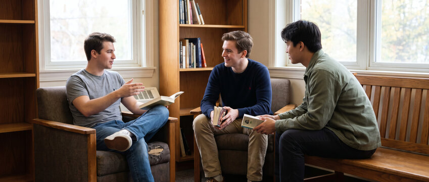 Three young men hold books, discussing casually in a cozy indoor space with wooden shelves and windows.