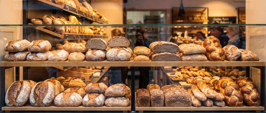 Various types of freshly baked breads are neatly arranged on shelves in a bakery display case, with customers in the background.