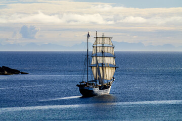 Traditional sailing ship cruises off Nusfjord, with the mountain silhouettes of the Lofoten Islands visible on the horizon. © EKH-Pictures