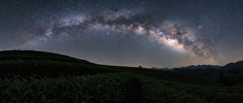 A starry night captures the Milky Way arching over a dark, vegetated hilly landscape with distant mountain silhouettes.
