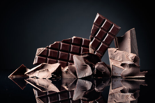 A broken chocolate bar and pieces of dark chocolate on a black reflective background.