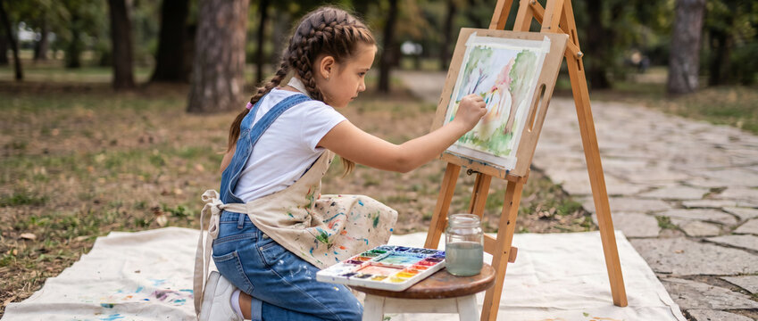 A young girl concentrates on painting with watercolors on an easel while sitting outdoors in a park, with art supplies nearby.