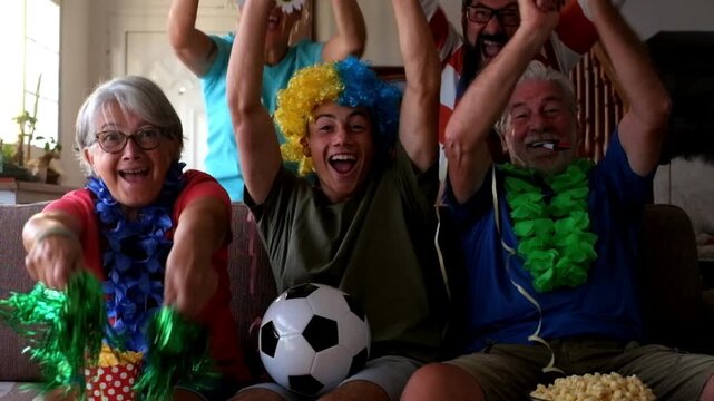 Enthusiastic multi-generational family wearing colorful wigs and accessories celebrating a goal while watching a soccer match on television, sitting on the sofa in the living room at home