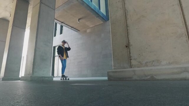 Low-angle view of a stylish young man skateboarding on a longboard through a modern concrete building, enjoying his urban lifestyle and freedom while riding towards the camera