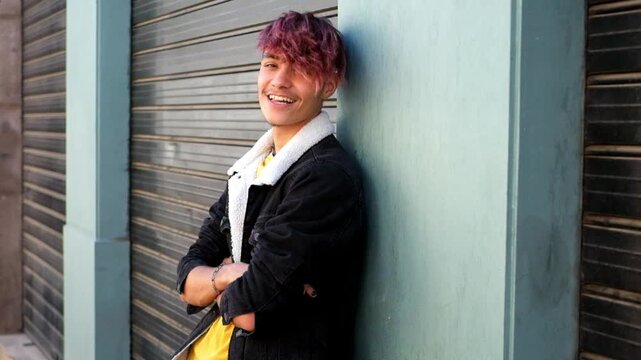 Stylish young adult man with purple dyed hair leaning against a wall outdoors, smiling and looking at the camera with confidence while posing with folded arms in an urban setting
