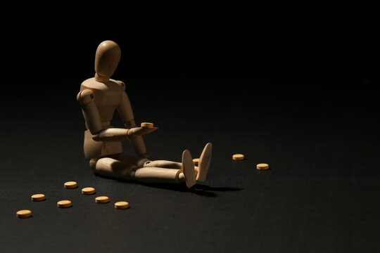 Wooden mannequin sitting on the floor holding a yellow pill surrounded by scattered medicine. Concept of depression, mental health, medical treatment or addiction.