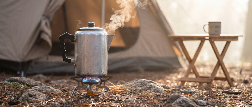 A metal coffee pot boils water on a portable camping stove, with a tent and small table in the outdoor campsite background.