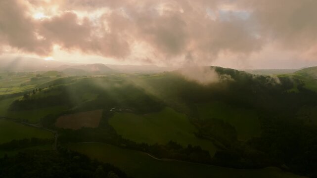 Aerial view of rolling green hills in Sao Miguel Island at sunset, with dramatic rays of light piercing through clouds over the pastoral landscape.