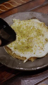 A hand pours clear syrup from a small black cup over a round kunafa topped with a creamy layer and crushed pistachios.