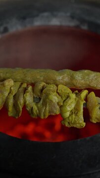 Marinated chicken strips and a long seekh kebab cook on metal skewers over glowing coals and flickering tandoor flames.