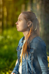 Side profile portrait of young woman with long red hair in sunlit forest, wearing denim jacket, exhaling subtle vapor suggesting vaping. Warm golden hour light, soft green bokeh, spring nature. © Viktoria