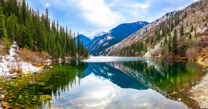 Lake Kolsai in spring. A beautiful spring mountain landscape in Kazakhstan. The blue lake is set against a backdrop of green spruce trees and still-snow-capped mountains.