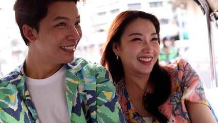 Fototapeta premium Happy young Asian couple wearing colorful shirts smiling and riding tuk tuk around city in Bangkok Thailand, romantic tourist man and woman enjoying vacation trip, local transit, tourism and journey