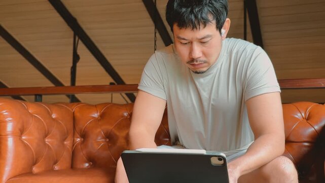 Concentrated Asian Man Using Digital Tablet with Stylus and Earbuds on Sofa