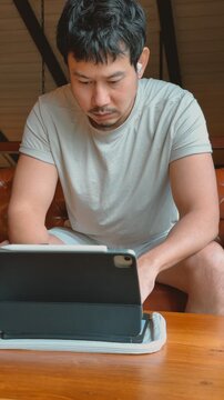 Asian man in casual wear intently using a digital tablet with wireless earbuds indoors.