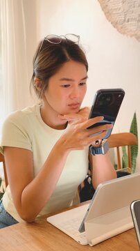 Young Asian woman intently using her smartphone at a wooden table with a tablet.