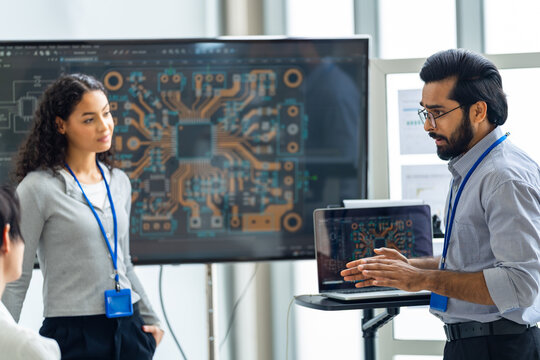 Bearded engineer with lanyard presents a circuit board design on a big screen to a smiling curly-haired colleague, gesturing over a laptop cart, an agentic AI and automation concept