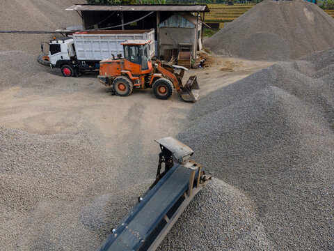 Heavy Machinery Orange Wheel Loader Near a Rock Crusher Machine with Conveyor Belt System and Aggregate Stockpiles Viewed from Above in Banyumas, Central Java, Indonesia.