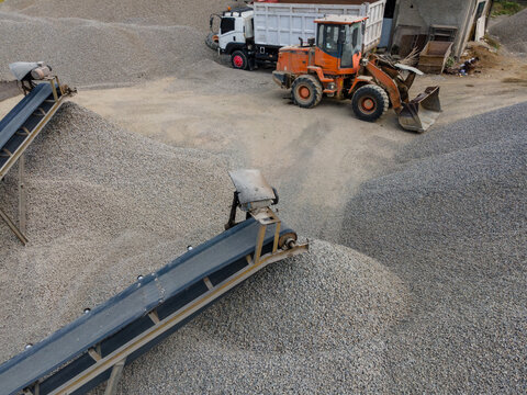 Heavy Machinery Orange Wheel Loader Near a Rock Crusher Machine with Conveyor Belt System and Aggregate Stockpiles Viewed from Above in Banyumas, Central Java, Indonesia.