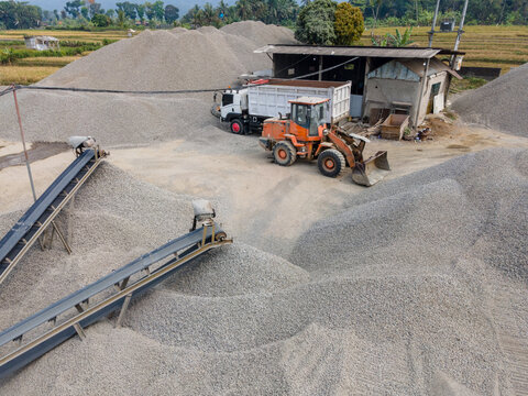 Heavy Machinery Orange Wheel Loader Near a Rock Crusher Machine with Conveyor Belt System and Aggregate Stockpiles Viewed from Above in Banyumas, Central Java, Indonesia.