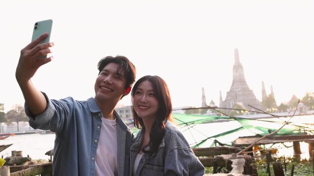 Happy young Asian couple taking selfie with smartphone, cheerful tourist man and woman enjoying city sightseeing trip near river temple landmark with Wat Arun, Bangkok, Thailand