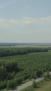 Wide aerial footage of dense green forest and a winding rural road with a car, under a blue sky in the Ural Mountains region, Russia, summer travel scenery