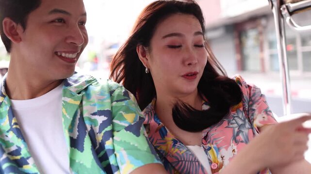 Happy young Asian couple wearing colorful shirts smiling and riding tuk tuk around city in Bangkok Thailand, romantic tourist man and woman enjoying vacation trip, local transit, tourism and journey