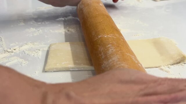 Close up of female hands using a wooden rolling pin to flatten pastry dough on a floured white table