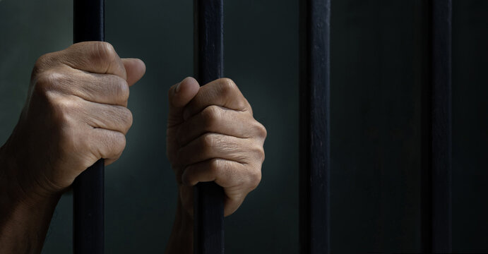 hands behind bars. Hand of convicted prisoner behind the cell bar inside jail for incarceration, criminal and limited freedom concept. Close-Up of Prison Inmate Gripping Jail Cell Bars Inside.
