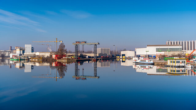 Industrial buildings, cranes, and boats reflecting in the calm water of a port under a clear blue sky