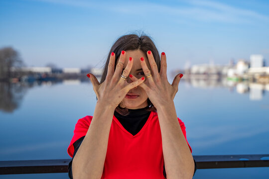 Woman covering face with hands and red nails, standing by a river under blue sky