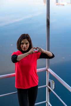 Woman in red shirt forming a heart shape with hands, standing by a lake railing