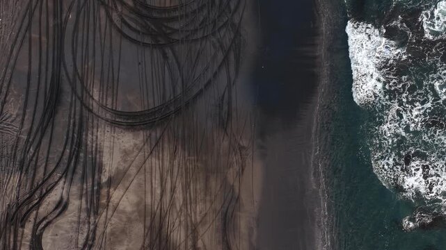 Aerial drone view shows tire tracks etched in black sand next to turquoise sea waves at Kelecung Beach, Tabanan, Bali, Indonesia  dramatic abstract patterns, textures and lines of the beach