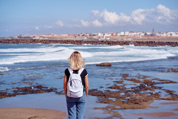woman with a backpack gazes at the ocean, intrigued by the coastal city views. The scene reflects themes of exploration, nature, and tranquility, capturing the adventurous spirit amid natural beauty. © Loginova