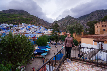 Obraz premium A woman stands on a balcony overlooking a vibrant town with blue-painted buildings, surrounded by mountains. The scene captures a bustling market square below, with people and stalls.