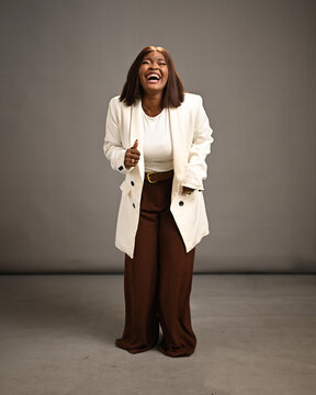 A confident professional woman laughs joyfully in a studio portrait wearing a white blazer and brown trousers, conveying happiness, charisma and approachable leadership for business, fashion and lifes
