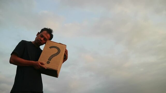 Man holding a cardboard box with a question mark, contemplating uncertainty against a cloudy sky.