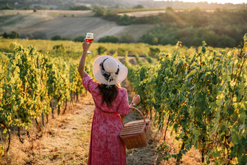 Obraz premium Woman raising glass of red wine in toast at sunset in vineyard