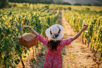 Obraz premium Woman standing with open arms holding wine glass and basket in vineyard