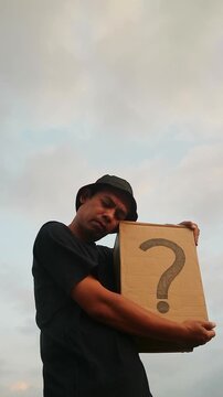 Man Embracing Cardboard Box with Question Mark Symbol Against a Cloudy Sky.