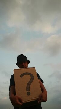 Person holding a cardboard box with a question mark against a cloudy sky, symbolizing mystery and uncertainty.