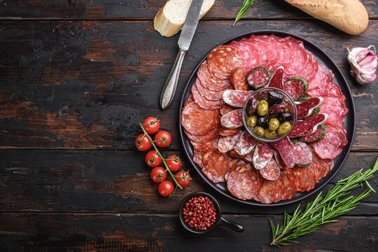 Round black ceramic plate filled with assorted sliced Spanish cured meats including chorizo salchichon fuets longaniza and lomo arranged in concentric circles around a glass bowl of green