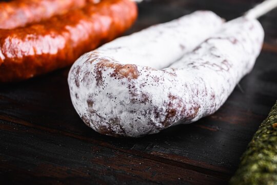 White powdery dry cured fuets arranged on dark rustic wooden surface beside reddish smoked sausages and green herb coated sausage under warm directional light