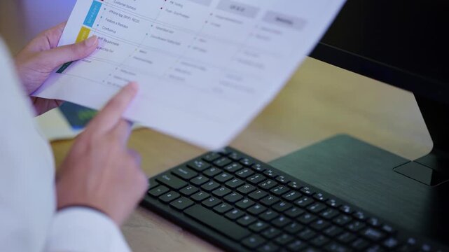 Close-up of hands holding a printed project report and checklist over a keyboard while reviewing work at a desk in a daylight workspace, while assessing the results, a physical AI concept