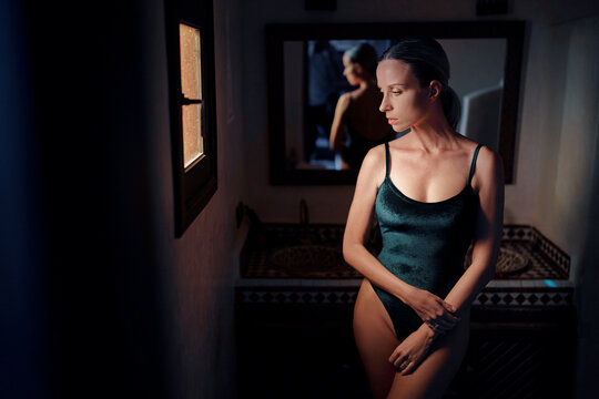A woman in a dark swimsuit stands in a dimly lit room, gazing out a window. The room features a mirror reflecting her image and a decorative sink in the background.