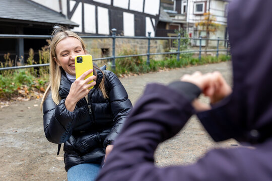 Smiling blonde woman photographs friend with smartphone during relaxed autumn walk in historic European town. Candid travel moment captures friendship, laughter, tourism, leisure, technology and joy