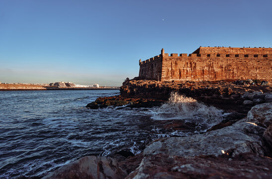 Stone fortress on a rocky shore overlooking the ocean under a bright, clear sky. Historic Stone Fortress by the Sea During Sunset